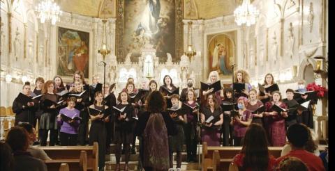 Le Choeur Maha, une trentaine de personnes non binaires et femme, chante dans une chapelle. Sarah Rossy, la directrice, a le dos à la caméra pendant qu'elle dirige le choeur.